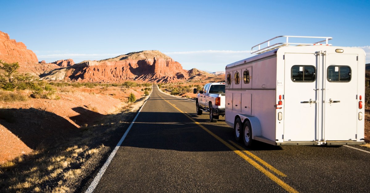 A white truck pulling a horse trailer down a secluded highway. The desert and mountainous terrain are visible on all sides.