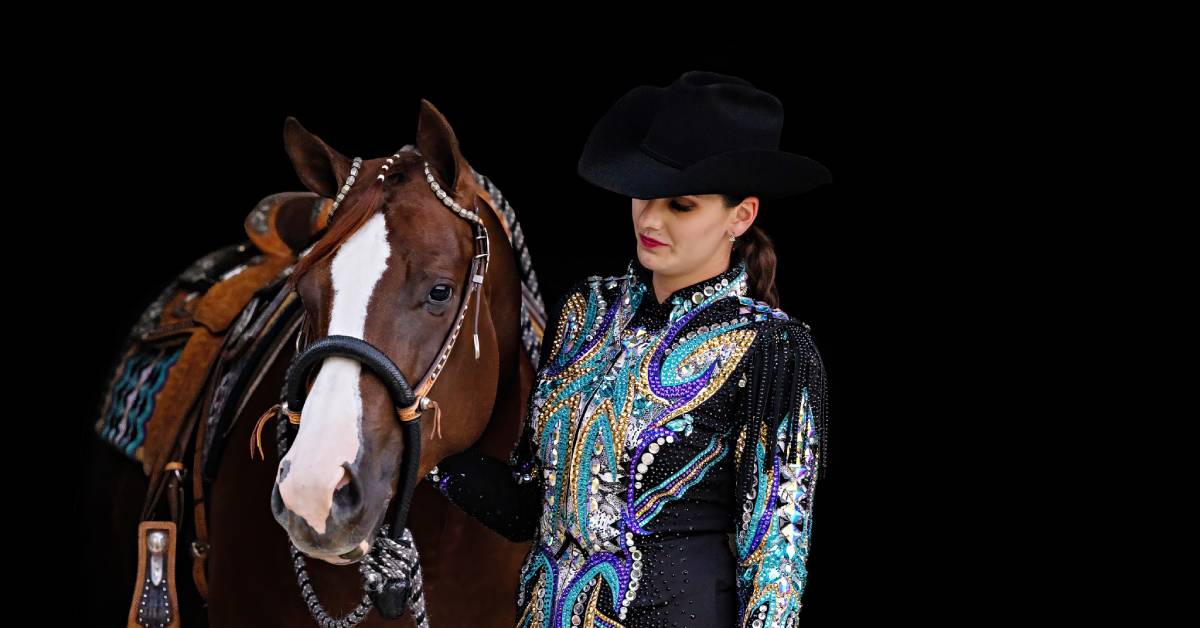 A woman stares at her horse and stands beside it. She's wearing a colorful show outfit with lots of sequins and beading.