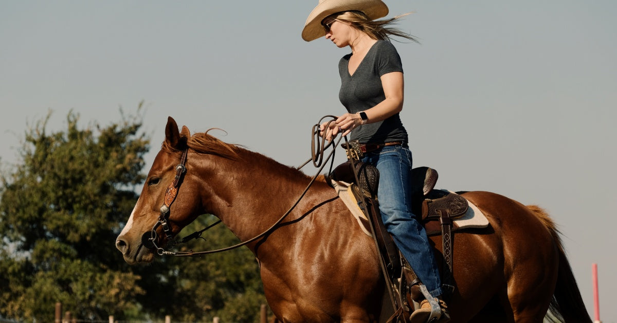 A blonde, white woman wearing a cowboy hat rides a brown horse. The woman sits on a saddle as she rides.