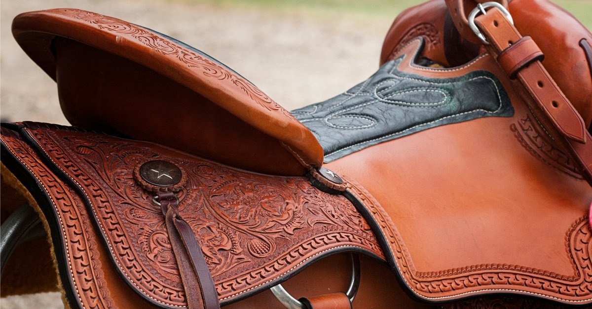 A brown leather Western saddle lays on top of a wood post. The saddle has intricate details woven into the leather.